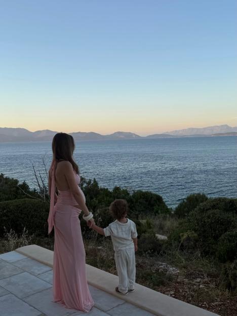 Lucy Watson, pregnant, holding hands with a child and looking out at the sea and mountains.