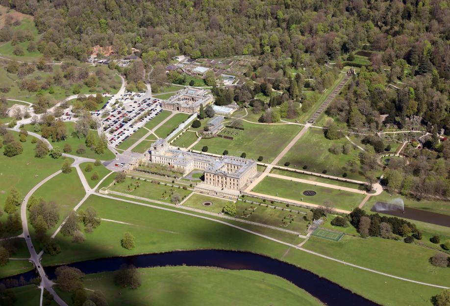 Aerial view of Chatsworth House Estate, Derbyshire, with a river flowing through lush green grounds, surrounded by woodlands.