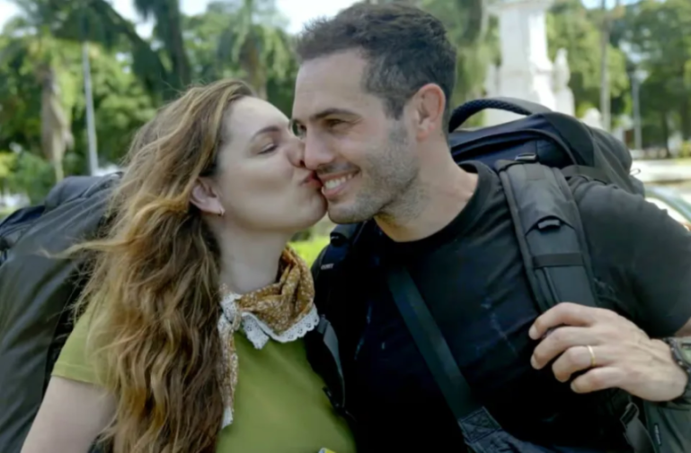 A woman kisses a man on the cheek while they both wear backpacks.