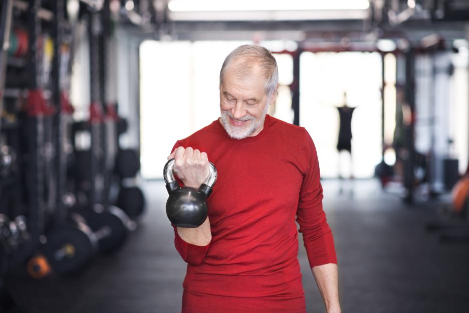 A senior man with a white beard, wearing a red long-sleeved shirt, smiles while curling a black kettlebell with his right arm in a gym.