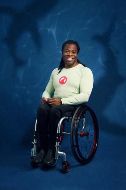 Ade Adepitan smiling in a wheelchair against a blue background with light ripples.