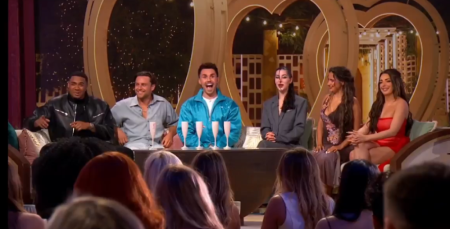 Six contestants, three men and three women, sit on a stage with champagne glasses in front of them, looking at an audience.