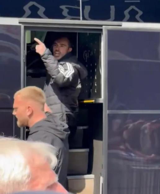 A man in a black puffer jacket points from the open door of a bus.