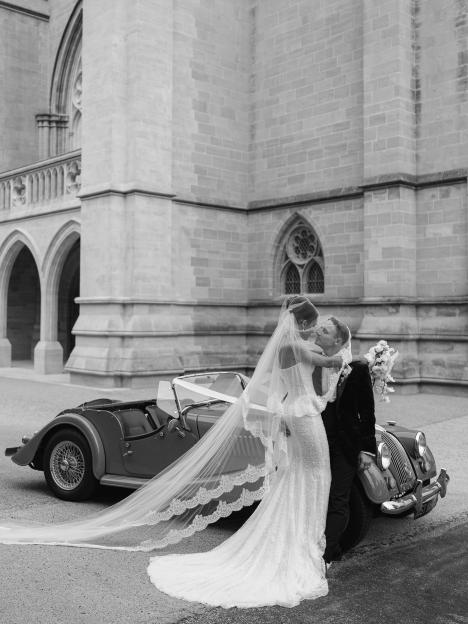 Natalie and Jack Visick embracing beside a vintage convertible car on their wedding day.