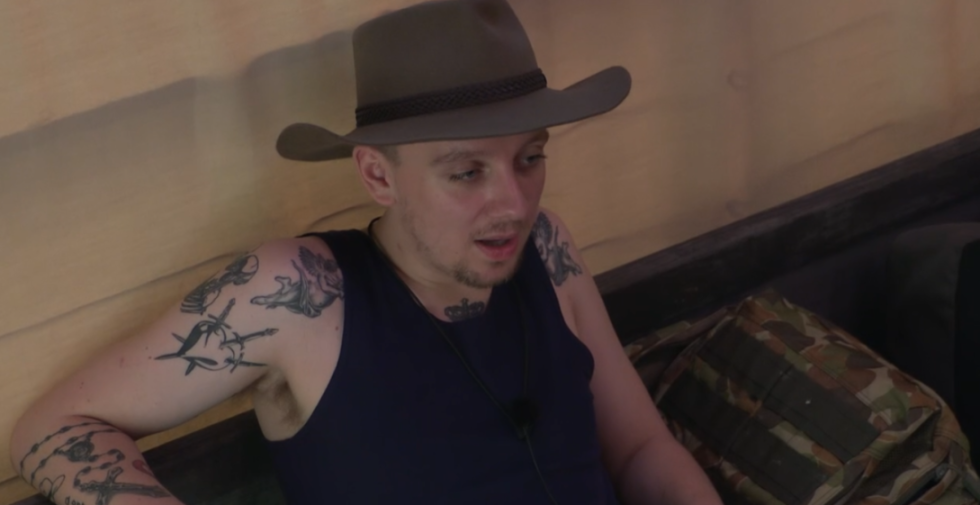 A man in a fedora, blue tank top, and tattoos on his arms and chest, looking left and talking.