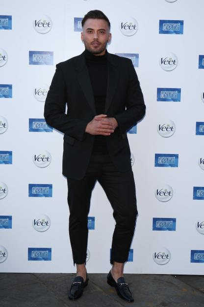 A man in a black suit with a black turtleneck and black loafers posing against a white backdrop covered in "Veet" and "The Only Way Is Essex" logos.
