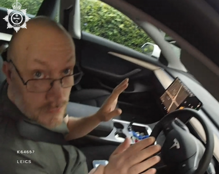 A man with glasses and a beard in a car with a Tesla steering wheel.