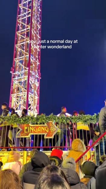 People queuing for a ride at a winter wonderland, with a large, lit-up tower in the background and an "Eingang" (entrance) sign.