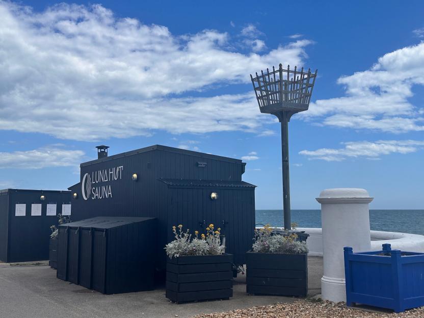The Luna Hut Sauna, a black building with white lettering, sits next to the sea with a tall beacon on the right.