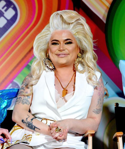 Baga Chipz, wearing heavy makeup, a blonde wig, and a white vest, sits and smiles at RuPaul's DragCon.