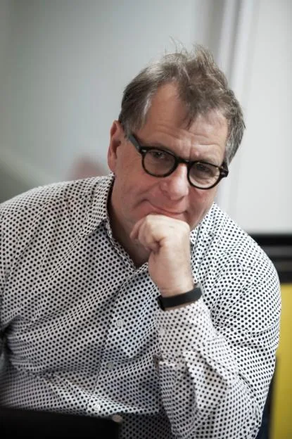 Portrait of a middle-aged man with glasses wearing a black and white polka-dot shirt, resting his chin on his hand.