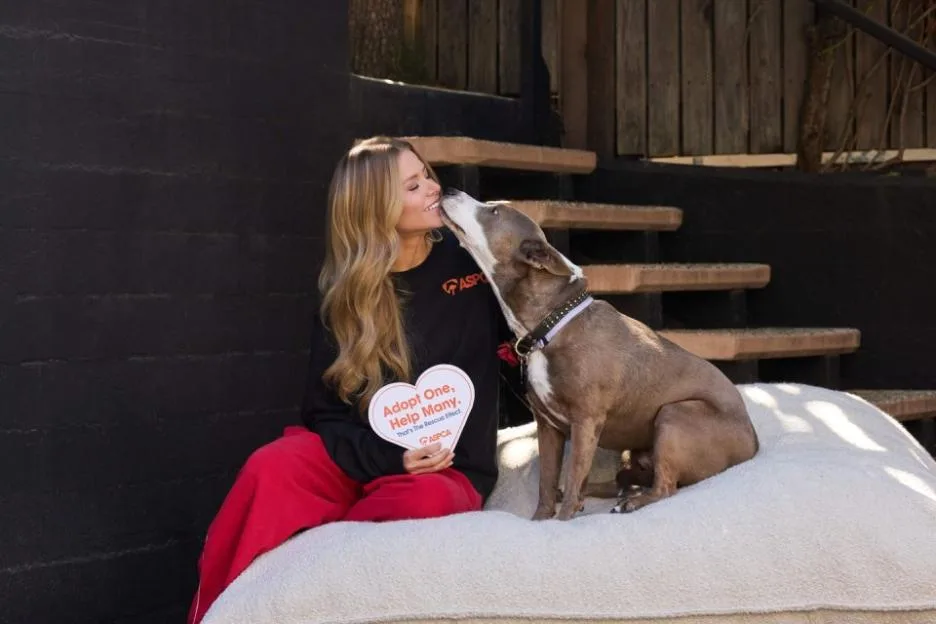 Ariana Madix sits on a cushion and holds a heart-shaped ASPCA sign while a dog nuzzles her cheek.