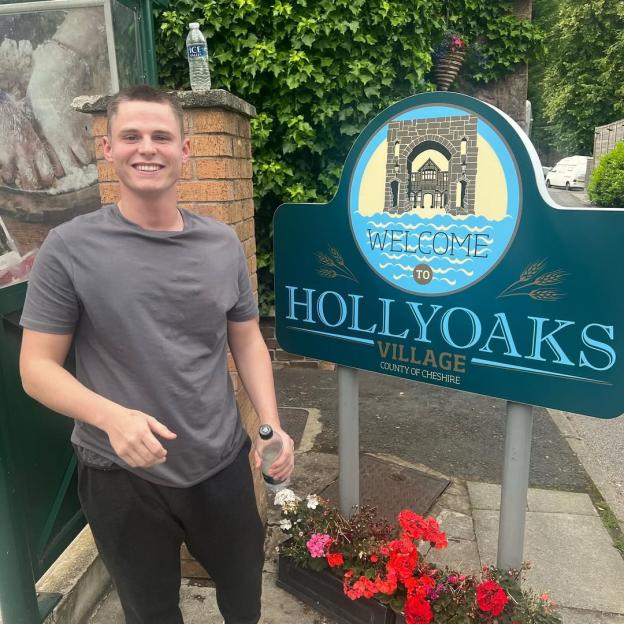 Billy Winter smiling next to a "Welcome to Hollyoaks Village" sign.