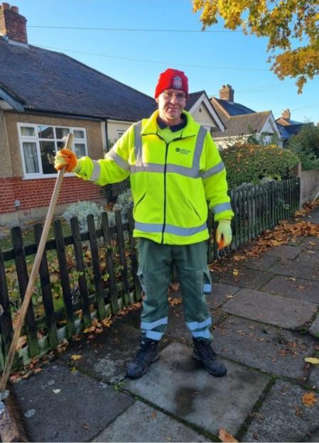Wayne Broadhurst in a bright yellow jacket, green pants, red beanie, and gloves, holding a broom.