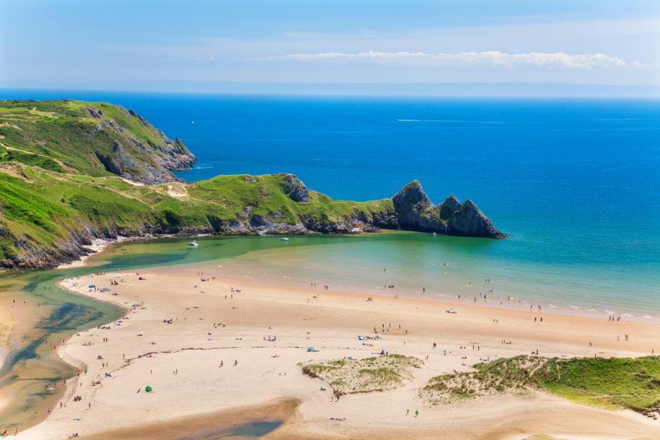 Three Cliffs Bay in Gower, Wales, UK, with people on the sandy beach and in the water.