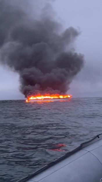 A 60-foot motor boat engulfed in flames, with thick black smoke billowing into the gray sky, seen from another boat in choppy dark water.