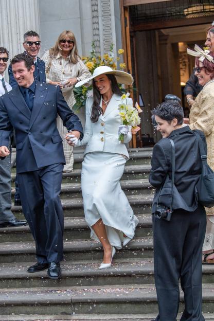 Dua Lipa and Callum Turner holding hands and smiling as they walk down steps covered in confetti after their wedding.