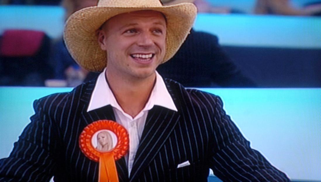 Richard Newman in a pinstripe suit, white shirt, and straw hat, wearing an orange rosette with a woman's picture.