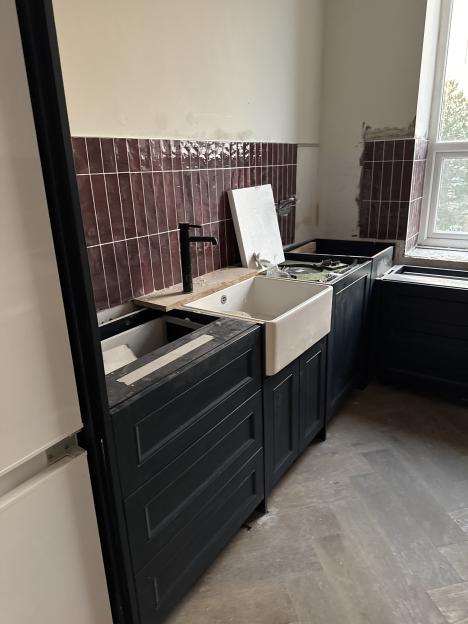 A partially installed kitchen with a farmhouse sink, new dark cabinets, and reddish-brown tiled walls.