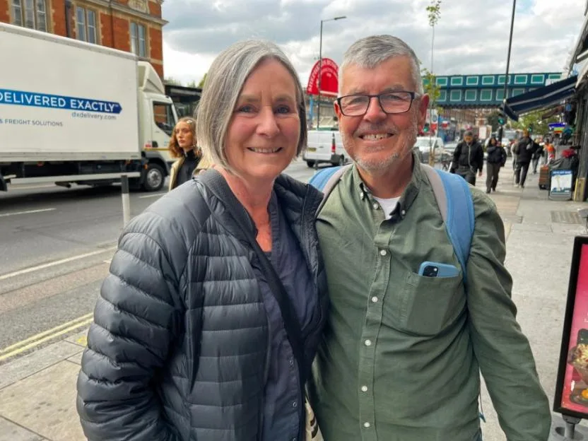 An older couple stands on the pavement of Uxbridge Road in London, with a lorry and other pedestrians visible in the background.