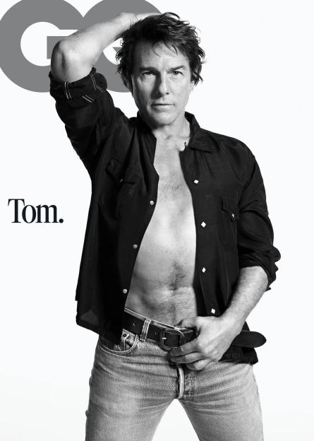 Tom Cruise posing for GQ Magazine, wearing an unbuttoned shirt and jeans, with "Tom." printed on the left side of the image.