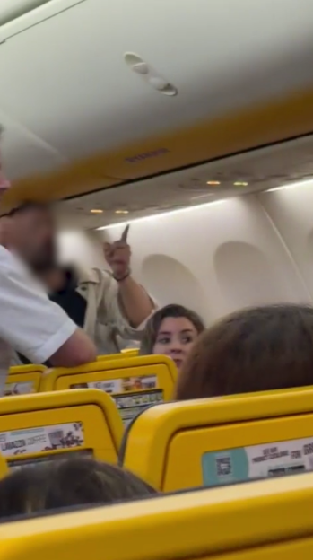 A man on an airplane points his finger aggressively while a woman behind him looks on.