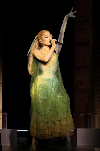 Ariana Grande performing onstage in a layered green dress with sheer sleeves that extend into gloves, holding a microphone with one arm raised.