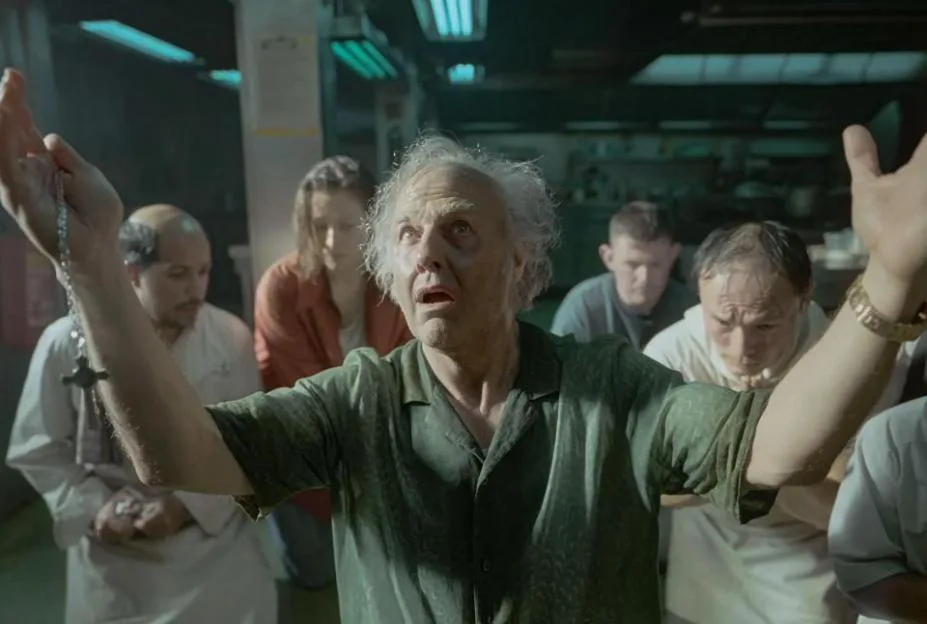An elderly man with wild white hair and a green shirt holds up a rosary and raises his hands in supplication among a group of distressed people.