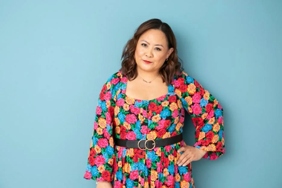 Michelle Agbulos, author of "Shine Baby Shine," stands against a blue background, wearing a vibrant floral dress and a black belt.