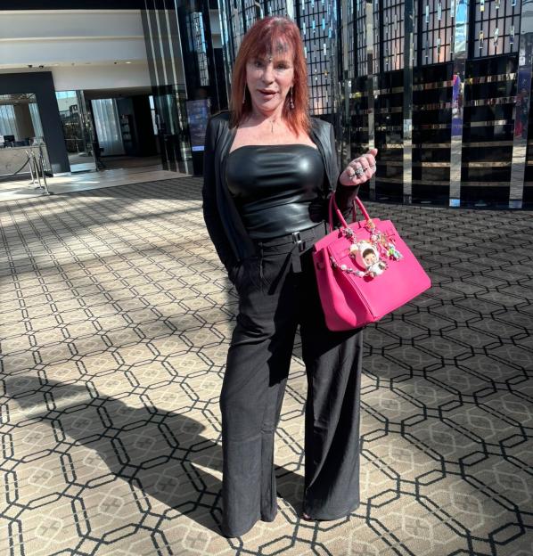 A woman with red hair, wearing a black leather top and black pants, holding a bright pink handbag.