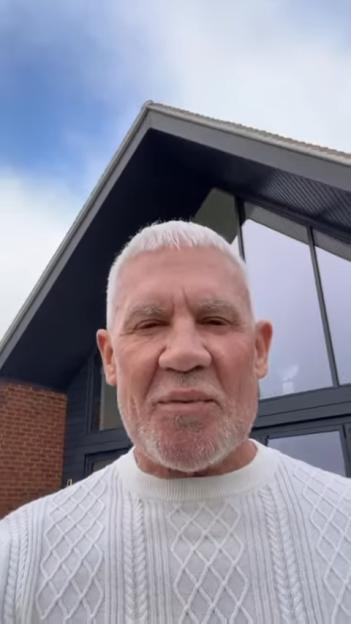 Wayne Lineker, with white hair and beard, wearing a white cable-knit sweater, standing in front of a modern building.