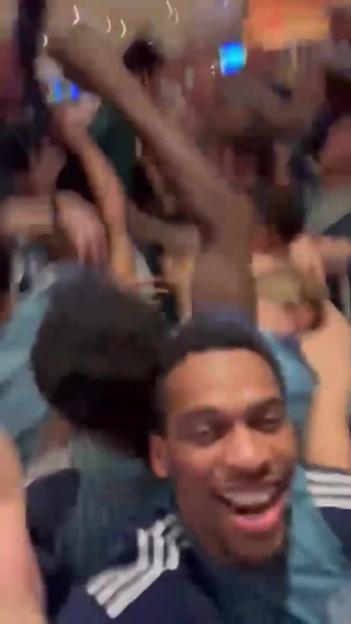 Man smiling at the camera amidst a crowd, with hands raised in the background.