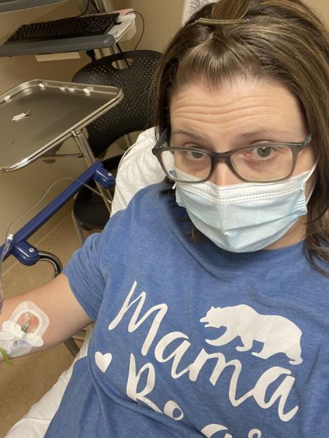 A woman wearing a face mask and a blue t-shirt with "Mama Bear" written on it, sits with an IV in her arm.