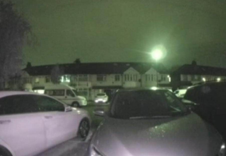 Night vision camera footage showing two cars parked in front of a row of houses, with a bright light in the sky.