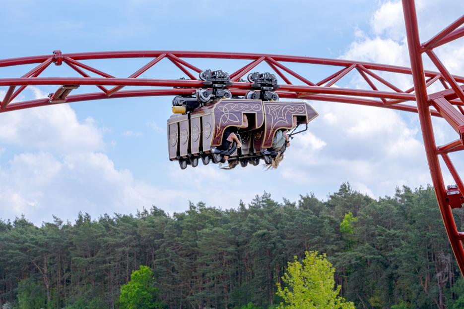 People upside down in a maroon roller coaster car on a red track with a forest in the background.