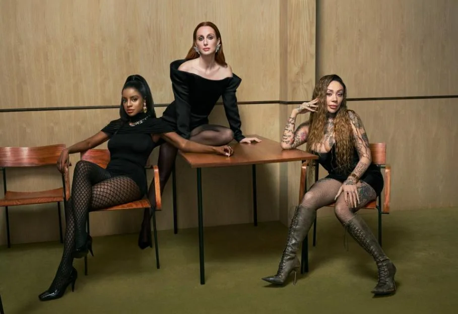 Three women in black dresses and stockings posing around a table.
