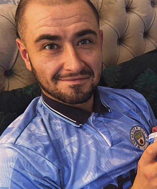 A smiling man wearing a light blue Manchester City F.C. shirt with a dark blue collar.
