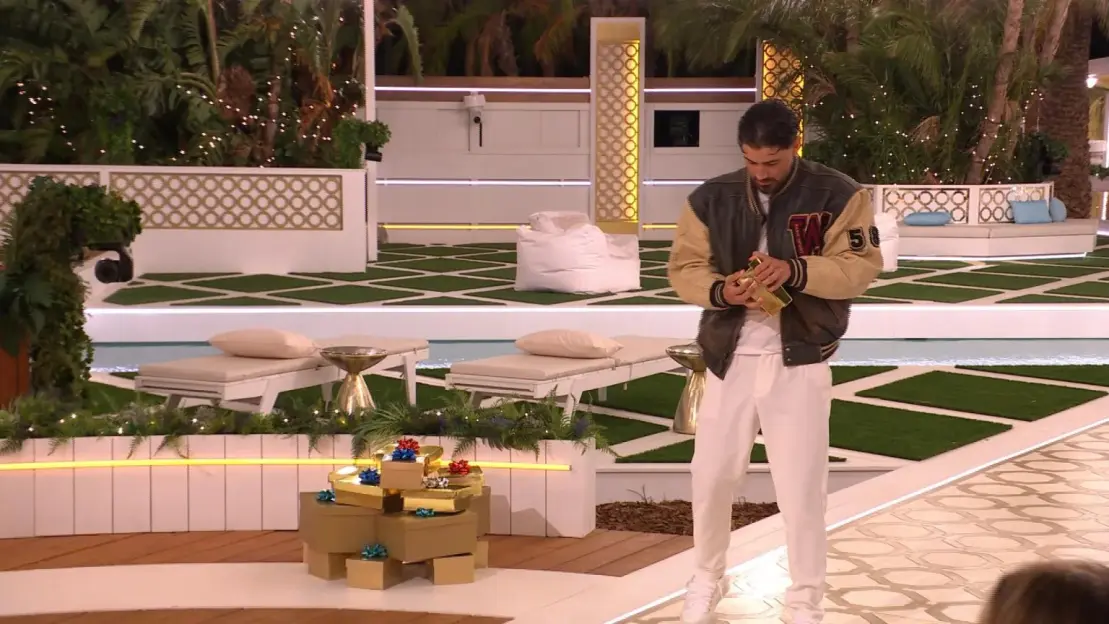 A man in a letterman jacket holds a golden bottle next to a stack of golden gift boxes.