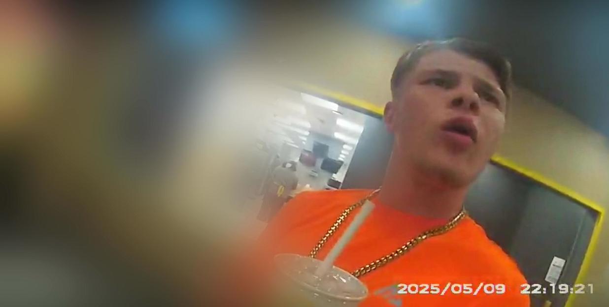Sonny Boyes wearing an orange shirt and gold chain, holding a drink and looking up, being arrested in a McDonald's.