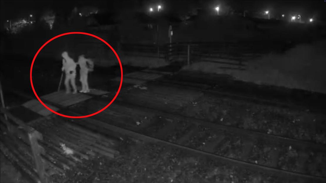 CCTV footage showing two individuals, one with a walking stick, crossing uneven railway tracks at night.