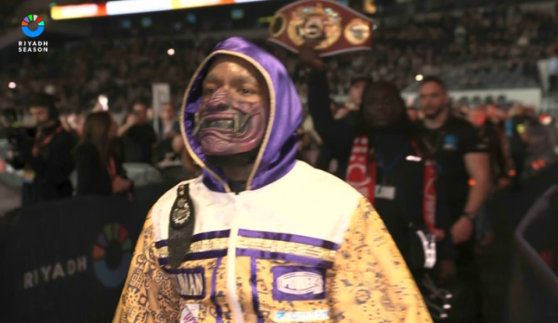 A boxer in a hooded robe and a face mask walking out for a fight.