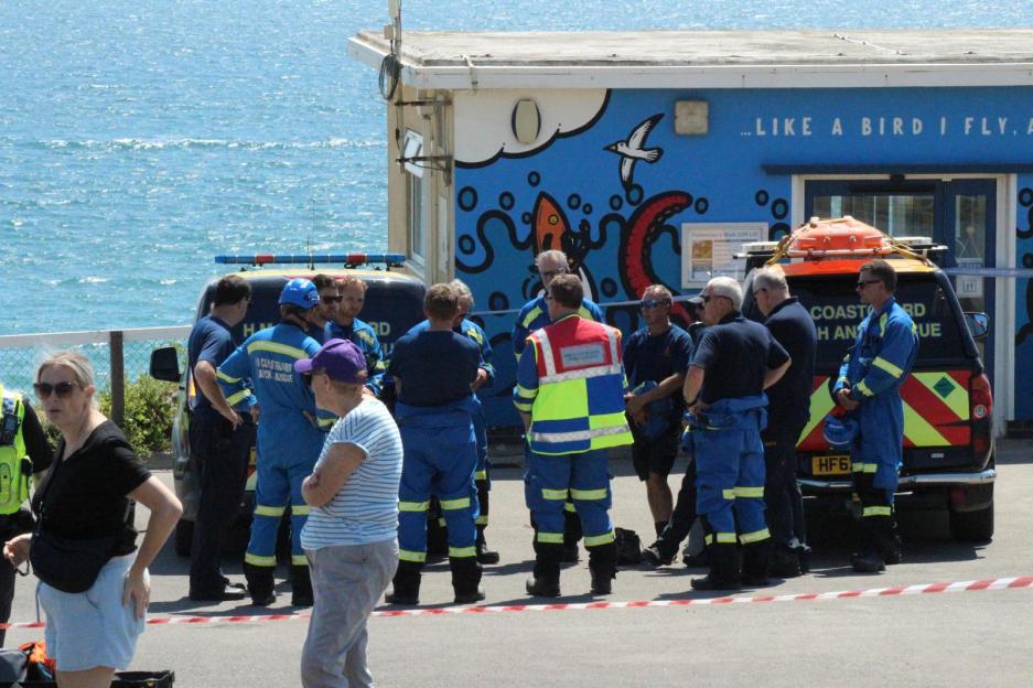Rescue workers gather near a building with "Coastguard Beach Rescue" vehicles parked nearby.