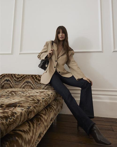 A model wearing a tan blazer, dark wash jeans, and brown boots sits on a patterned couch.