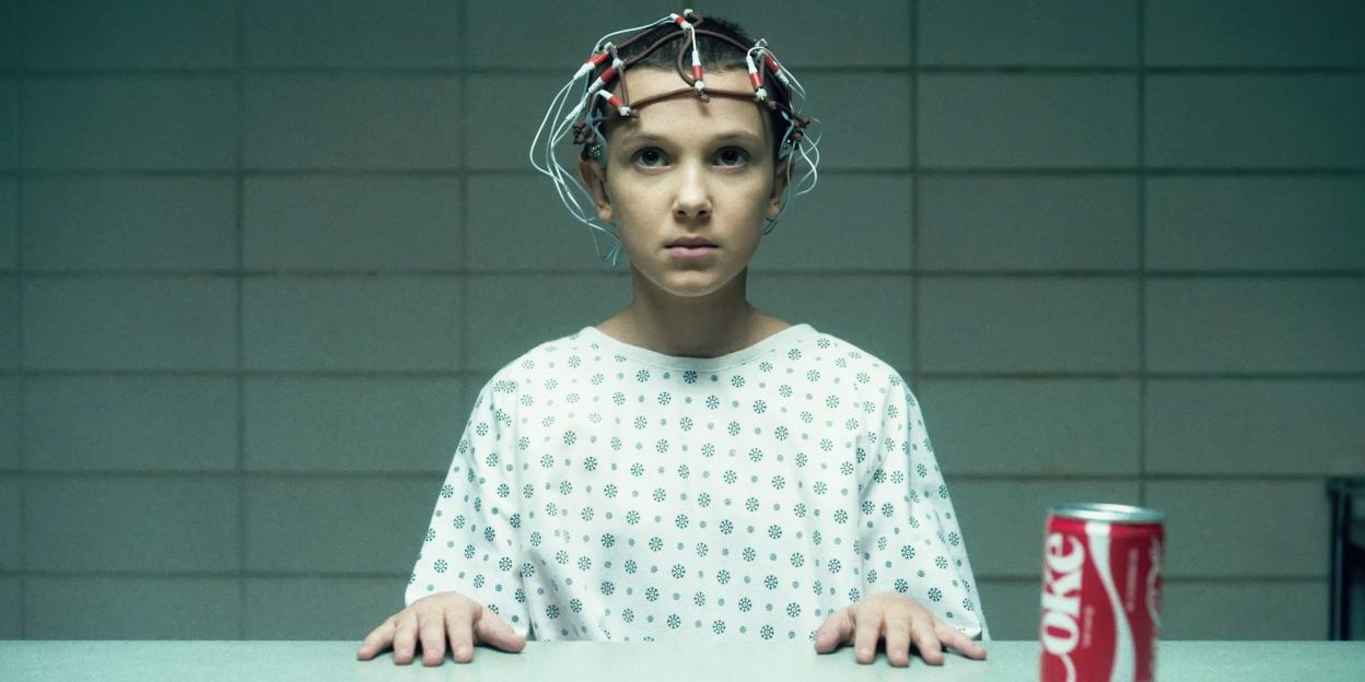 Millie Bobby Brown as Eleven in Stranger Things, wearing a patient gown with a device on her head, sitting at a table with a can of Coke.