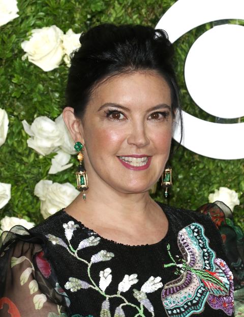 Phoebe Cates smiling in a black sequined top with butterfly embroidery, against a backdrop of green foliage and white roses.