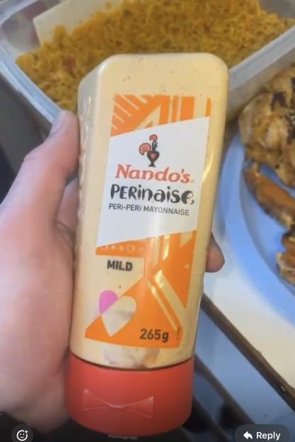 Hand holding a bottle of Nando's PERinaise, Mild peri-peri mayonnaise (265g), with food in containers in the background.