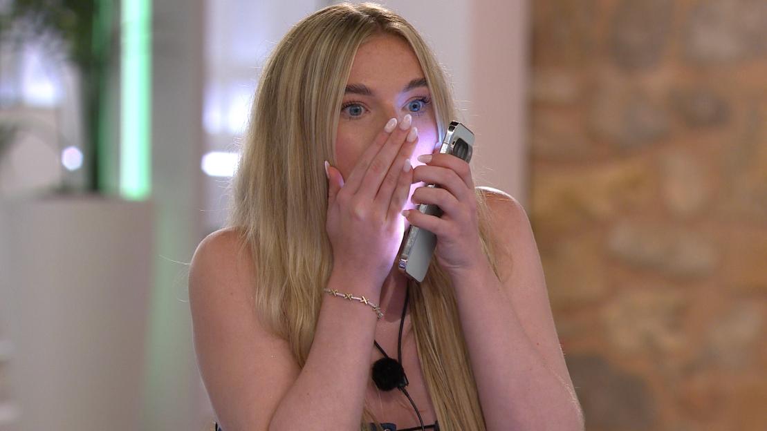 Robyn from Love Island reacting with surprise, covering her mouth with one hand while holding a phone close to her face.