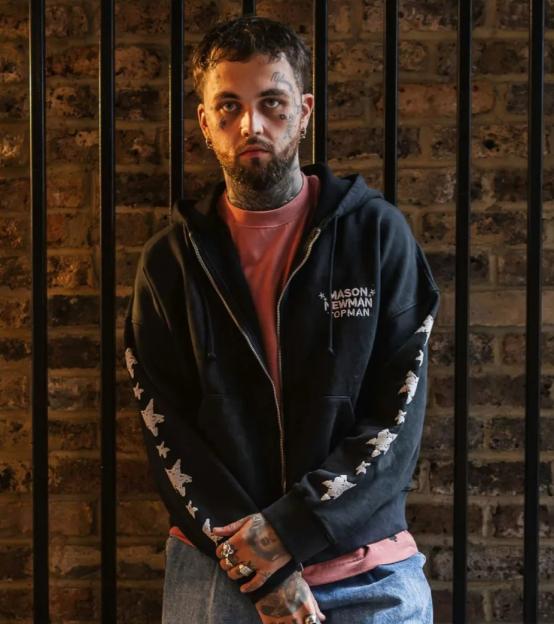 A man with face tattoos wearing a black hoodie with "MASON NEWMAN TOPMAN" and white star designs on the sleeves, a salmon-colored shirt underneath, and blue jeans.