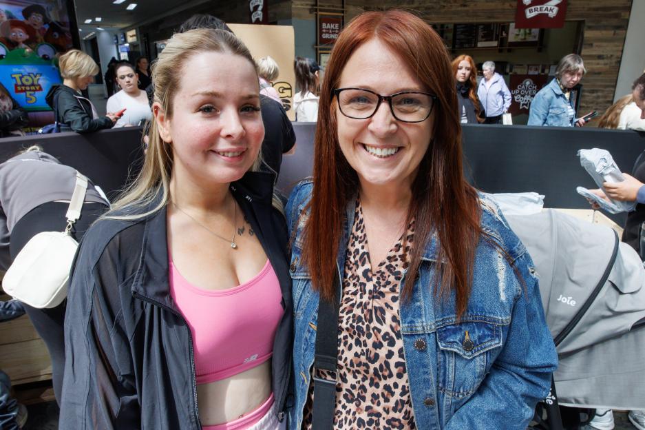 Two women smiling at the camera, one in a pink sports bra and black jacket, and the other in a leopard print top and denim jacket.