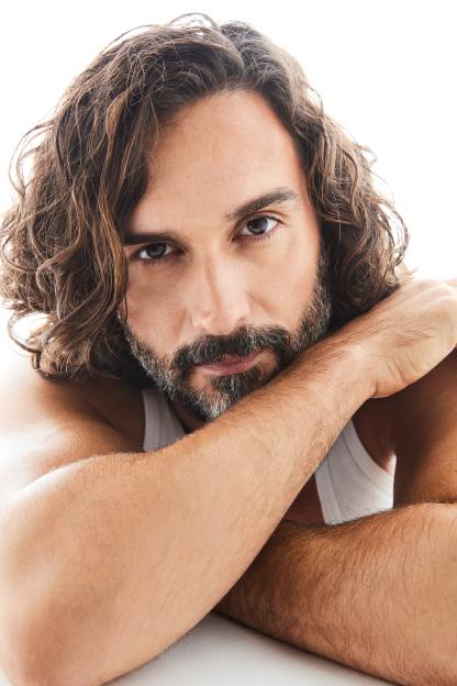 A man with long curly brown hair and a beard, wearing a white tank top, with his arms crossed in front of him.